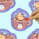 A hand with pink nail polish peels a die-cut sticker from a light blue background patterned with the same sticker design. The sticker features a cute hamster sleeping in a hot cocoa cup with a snowman on the cup, set against a light blue background.