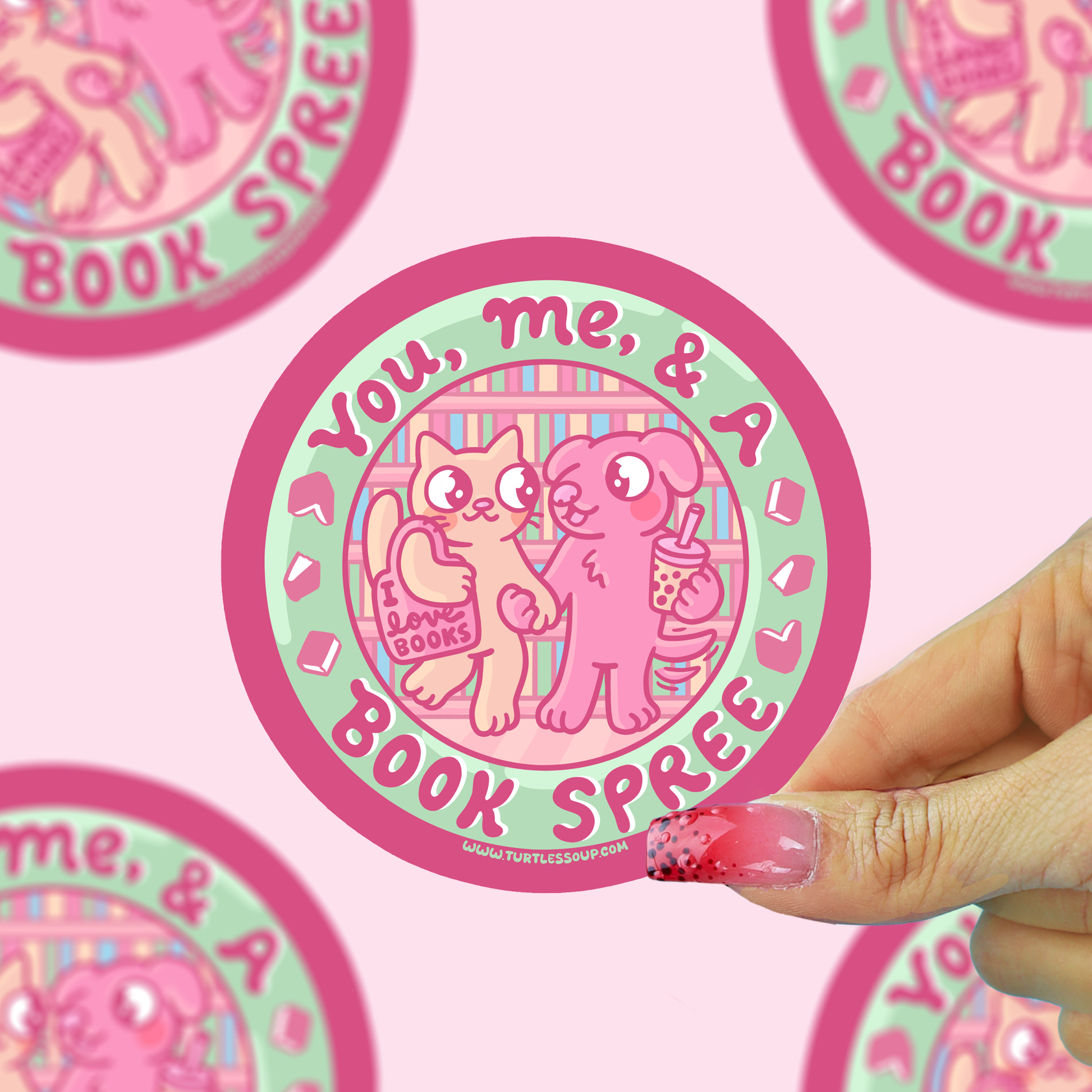 A hand with pink glittery nails holds a round sticker featuring a pink cat and dog character with the text 'You, me, & a BOOK SPREE'. The background is a light pink patterned surface with other partial stickers visible.