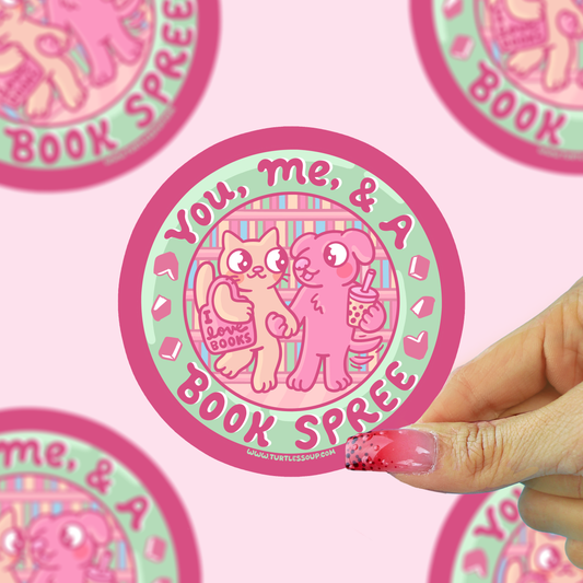 A hand with pink glittery nails holds a round sticker featuring a pink cat and dog character with the text 'You, me, & a BOOK SPREE'. The background is a light pink patterned surface with other partial stickers visible.