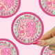 A hand with pink glittery nails holds a round sticker featuring a pink cat and dog character with the text 'You, me, & a BOOK SPREE'. The background is a light pink patterned surface with other partial stickers visible.