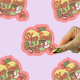 Sticker with 'Shop Small' design on a pink background, with a hand holding a gold marker.