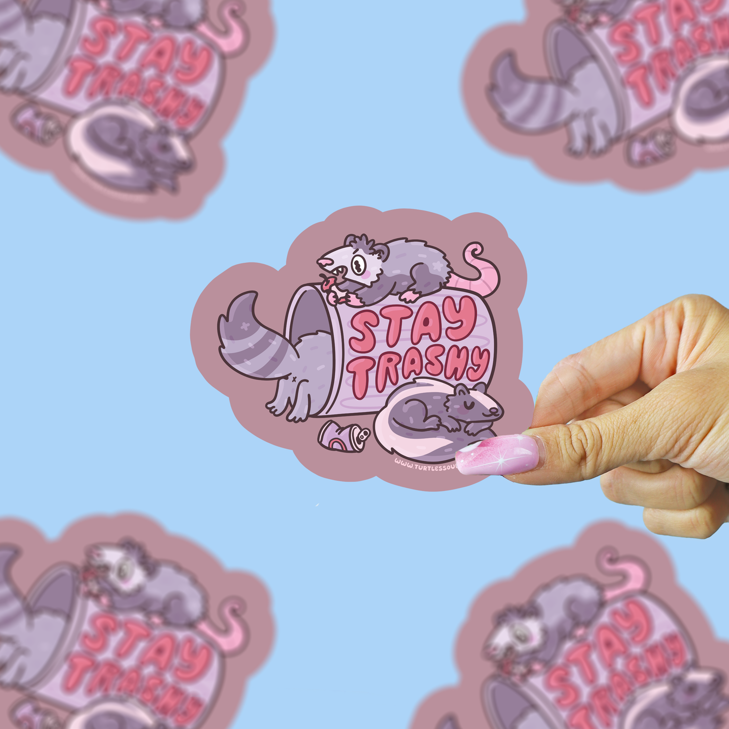 Sticker with a raccoon illustration and 'Stay Trashy' text, held by a hand against a blue background.