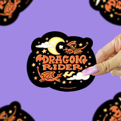 A hand with pink nail polish holds a black die-cut sticker featuring orange and yellow dragon-themed artwork and the words “DRAGON RIDER.” The background is lavender purple with several blurred copies of the same sticker around the edges.