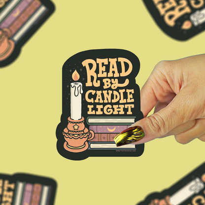 A hand with gold and green nail polish holds a die-cut sticker that reads 'READ BY CANDLE LIGHT' with an illustration of a candle and books. The background is a pale yellow with several blurred copies of the same sticker.