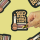 A hand with gold and green nail polish holds a die-cut sticker that reads 'READ BY CANDLE LIGHT' with an illustration of a candle and books. The background is a pale yellow with several blurred copies of the same sticker.