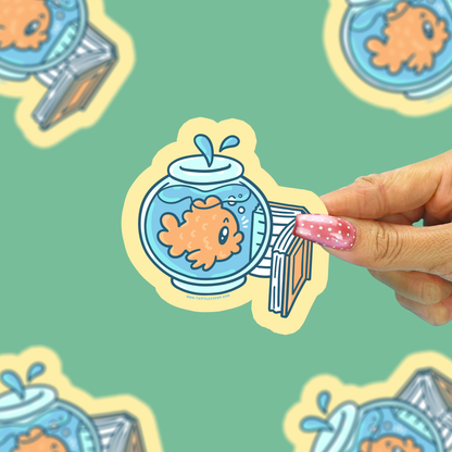 A hand with pink glitter nail polish holds a die-cut sticker featuring a cartoon goldfish in a blue fishbowl with a striped diving board. The background is a mint green patterned surface with other blurred stickers.