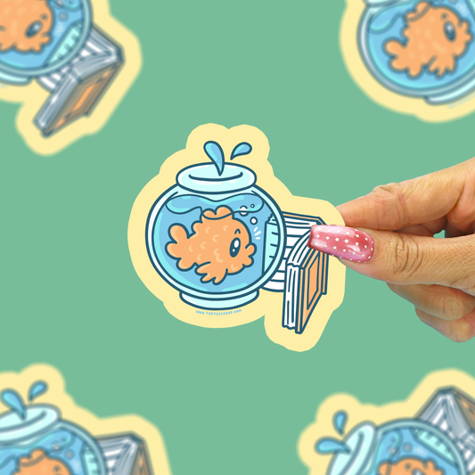 A hand with pink glitter nail polish holds a die-cut sticker featuring a cartoon goldfish in a blue fishbowl with a striped diving board. The background is a mint green patterned surface with other blurred stickers.
