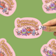 A hand holds a pink die-cut sticker featuring cartoon animals and the text 'Community is Resistance!' against a green background with blurred duplicate sticker previews.