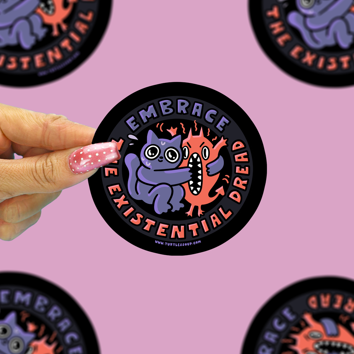 A hand with pink glitter nail polish holds a round sticker featuring a purple monster hugging an orange fire creature. The sticker has a black border with the text 'EMBRACE THE EXISTENTIAL DREAD'.