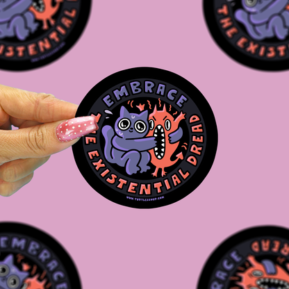 A hand with pink glitter nail polish holds a round sticker featuring a purple monster hugging an orange fire creature. The sticker has a black border with the text 'EMBRACE THE EXISTENTIAL DREAD'.
