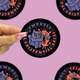 A hand with pink glitter nail polish holds a round sticker featuring a purple monster hugging an orange fire creature. The sticker has a black border with the text 'EMBRACE THE EXISTENTIAL DREAD'.