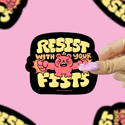 A hand holds a die-cut sticker featuring a pink cartoon bear with clenched fists and the text 'RESIST WITH YOUR FISTS' in yellow on a black background. The sticker is centered against a pink backdrop with several blurred copies of the same sticker around the edges.
