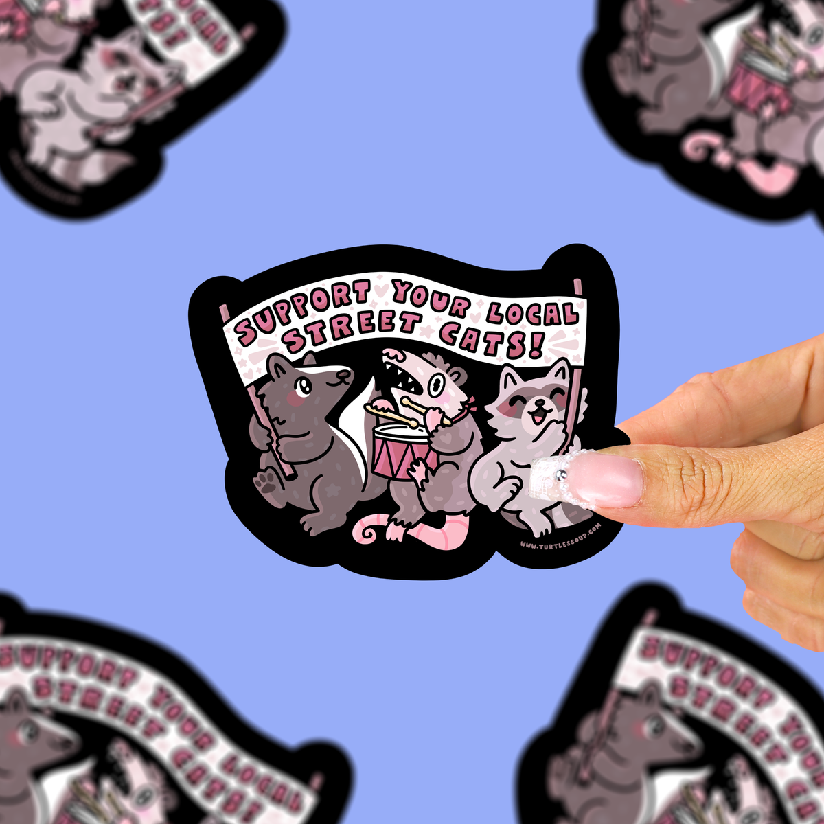 A hand holds a die-cut sticker featuring three cartoon street cats under a banner that reads 'SUPPORT YOUR LOCAL STREET CATS!' against a light blue background with blurred duplicate sticker images around the edges.