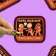 A hand with green nail polish holds a square sticker featuring a retro-style illustration with the text 'CATS AGAINST CAT-CALLING' and a stop sign. The background is a solid lavender purple with several blurred copies of the sticker.