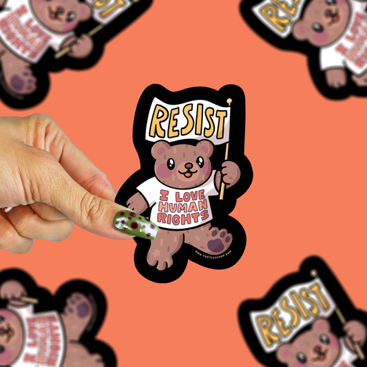 A hand holds a die-cut sticker featuring a cartoon teddy bear holding a sign that says 'RESIST' and wearing a shirt that reads 'I LOVE HUMAN RIGHTS'. The background is a solid orange color with blurred duplicate images of the sticker.