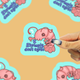A hand is applying clear nail polish to a die-cut sticker featuring a pink shrimp character with the text 'I Shrimply don't agree'. The background is a warm tan color with other copies of the sticker pattern visible.