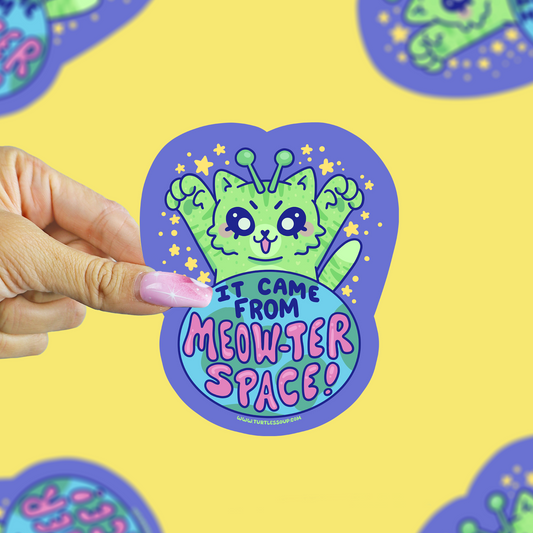 A hand with pink manicured nails holds a die-cut sticker featuring a cute green cartoon cat with alien antennae and the text 'IT CAME FROM MEOWTER SPACE!' on a bright yellow background with other blurred stickers.