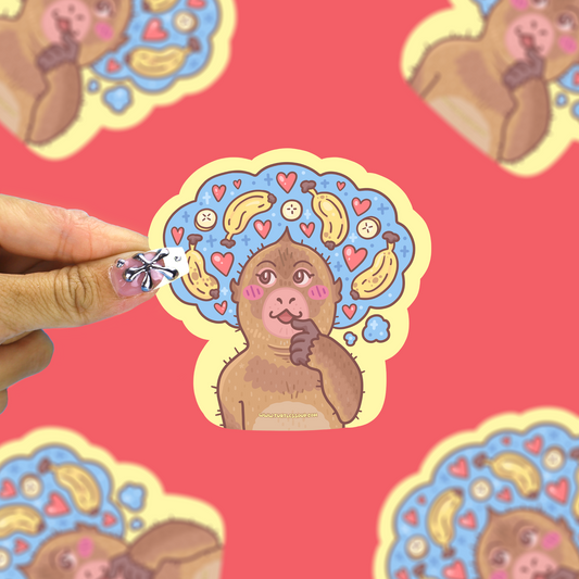 A hand holds a die-cut sticker featuring a cartoon monkey with a blue head full of bananas, hearts, and faces, set against a pink background with blurred duplicate stickers.