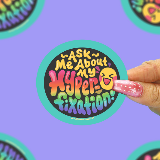 A hand with pink glittery nails holds a round sticker that reads 'ASK ME About My Hyper-fixation!' in colorful retro lettering on a black background with a turquoise border. The background is a solid lavender purple with blurred, out-of-focus versions of the same sticker.