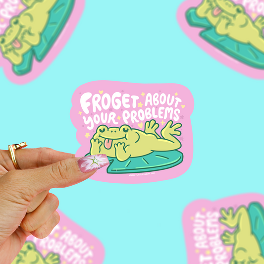 A hand holds a die-cut sticker featuring a cartoon frog lounging on a lily pad with the text 'FROGET ABOUT YOUR PROBLEMS' on a pink background. The sticker is shown against a light blue backdrop with several matching stickers scattered around.