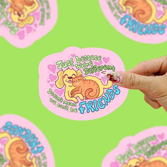 A hand holds a pink die-cut sticker featuring a cartoon dog and cat hugging with the text 'Just because we were different we couldn't be friends' on a bright green background with other blurred stickers.