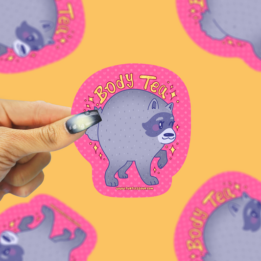 A hand with dark glittery nail polish holds a die-cut sticker featuring a cartoon gray raccoon with a pink polka-dot outline and the text 'Body Tea'. The background is a solid yellow surface with other identical stickers scattered around.