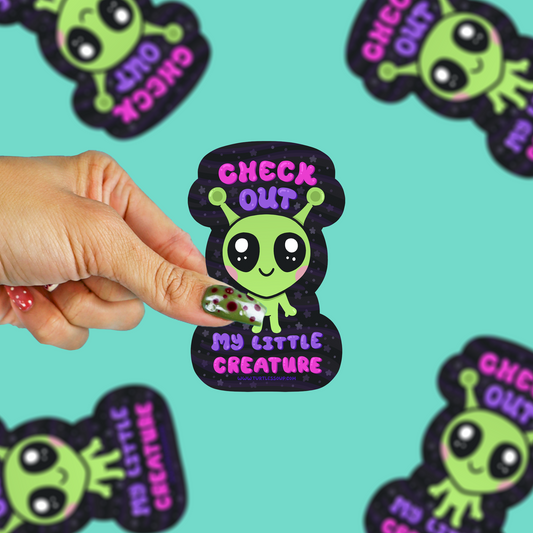A hand holds a small sticker featuring a cute green alien with large eyes and the text 'CHECK OUT MY LITTLE CREATURE' on a black glittery background. The sticker is shown against a bright aqua background with several matching stickers scattered around.