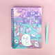 A spiral-bound notebook with a cute Halloween-themed illustration featuring a haunted house, ghosts, tombstones, bats, and a moon on a pink cover, shown on a pink background next to a mint green tweezers tool.
