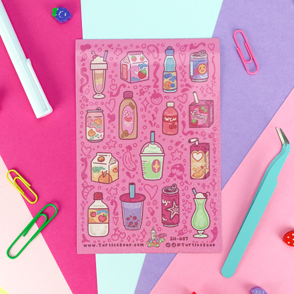 A pink sticker sheet featuring cute illustrations of various milk drinks, smoothies, and juice boxes, styled in a flat-lay on a geometric background of pink, purple, and light blue paper with office props.