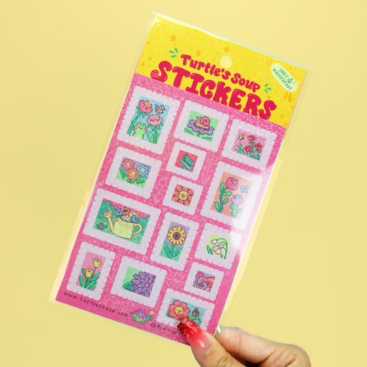 A hand holding a package of Turtle Soup Stickers against a solid pale yellow background. The package contains 12 colorful garden-themed stickers on a pink sheet with a yellow header card.