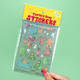 A hand with red manicured nails holds a package of 'Turtle's Soup Stickers' against a solid mint green background. The package contains various colorful cartoon stickers of monsters, ghosts, and creatures on a glittery teal backing card with a yellow header.