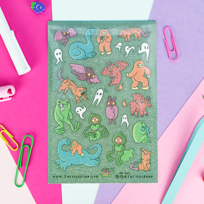 A flat-lay of a sticker sheet featuring various cute cartoon monsters and ghosts on a green background, surrounded by colorful paper clips and stationery on a pink, purple, and blue geometric background.