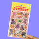 A hand holding a sheet of 'Turtle's Soup STICKERS' featuring cute, colorful cartoon cats in Halloween costumes on a pastel pink background with a yellow header card. The background of the photo is a solid lavender purple.