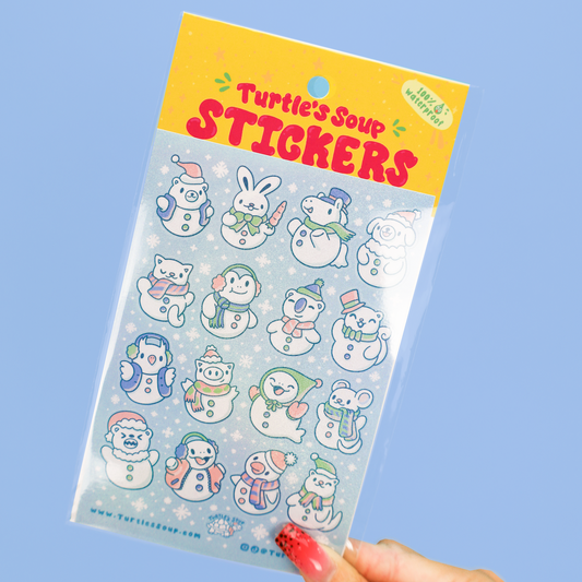 A hand holds a package of cute winter-themed stickers featuring snowman characters in various outfits, set against a solid light blue background.