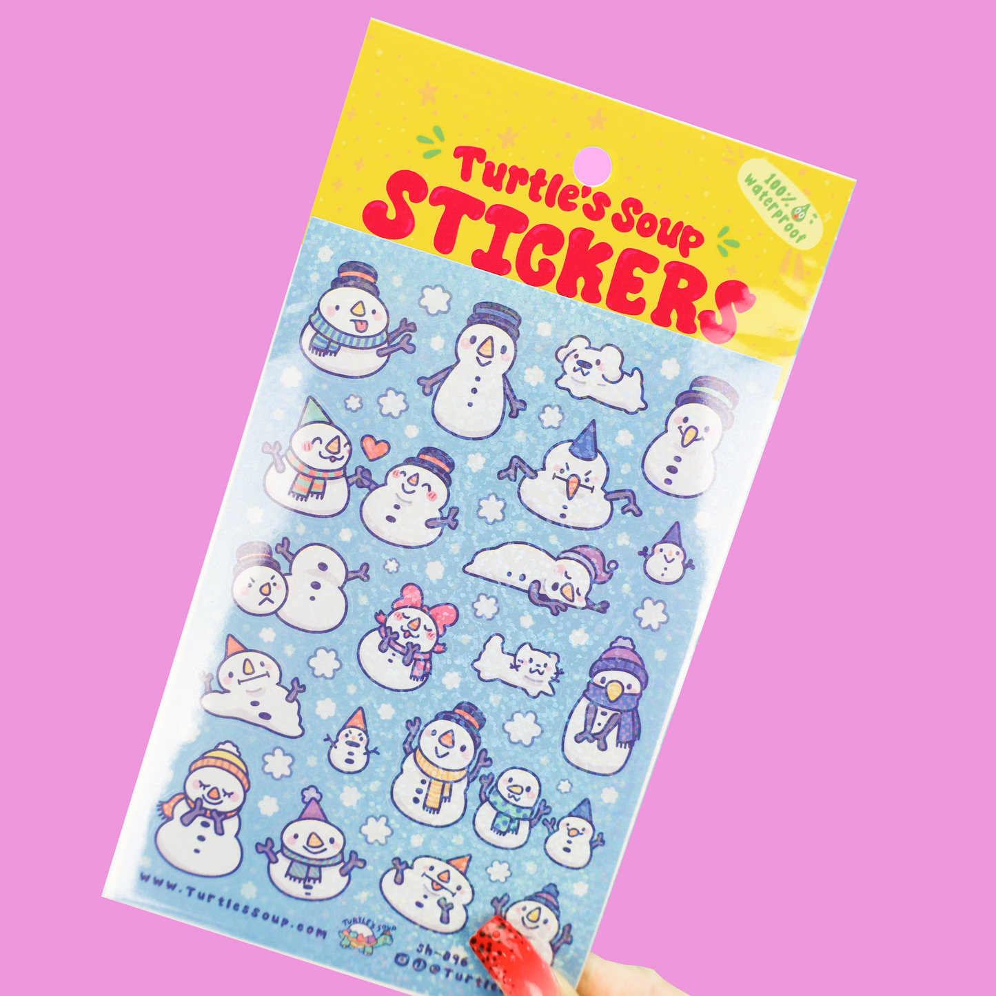 A hand holds a package of cute cartoon turtle soup stickers featuring snowmen and winter characters on a light blue background with a yellow header, set against a solid pink backdrop.