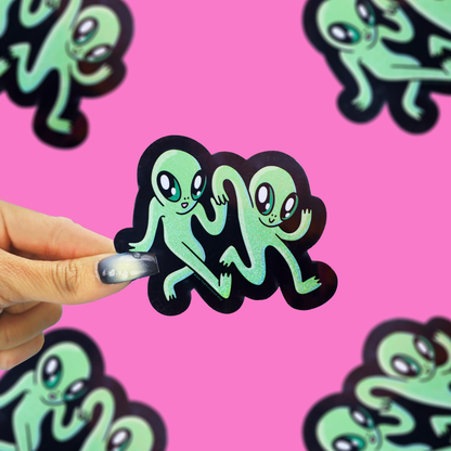 A hand holds a green glitter alien sticker with a black outline against a bright pink background with other matching alien stickers.