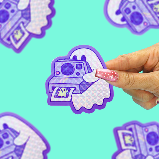 A hand with pink glittery nails holds a die-cut sticker featuring a purple retro instant camera and ghost illustration. The background is a solid mint green with other blurred versions of the sticker.