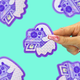 A hand with pink glittery nails holds a die-cut sticker featuring a purple retro instant camera and ghost illustration. The background is a solid mint green with other blurred versions of the sticker.