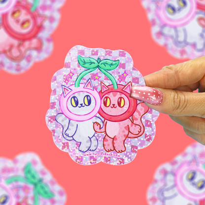 A hand with pink glitter nail polish holds a scalloped sticker featuring two cartoon cherry-like cats, one white and one pink, on a pink glitter background. The hand is positioned on the right side of the sticker, and other blurred stickers are visible in the background against a solid pink backdrop.
