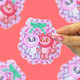 A hand with pink glitter nail polish holds a scalloped sticker featuring two cartoon cherry-like cats, one white and one pink, on a pink glitter background. The hand is positioned on the right side of the sticker, and other blurred stickers are visible in the background against a solid pink backdrop.