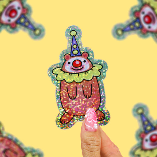 A hand with pink glittery nails holds a small, glittery clown sticker against a bright yellow background with other blurred clown stickers.