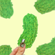 A hand with patterned nail art holds a green glittery pickle-shaped item against a pale yellow background with blurred pickle silhouettes.