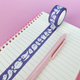 Purple washi tape with white cartoon ghosts, shown unrolled across a lined notebook next to a pink pen on a pink background.