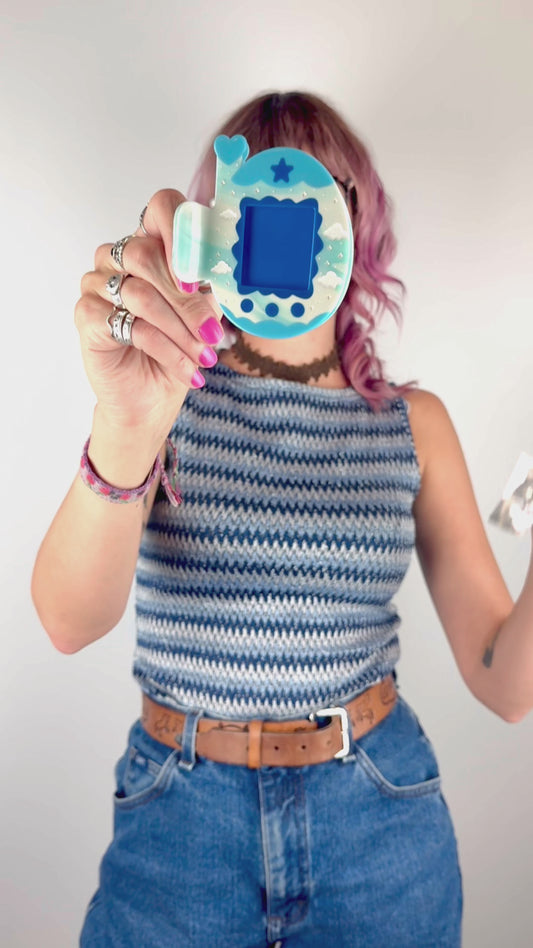 A close-up lifestyle shot of a person's hair secured with a light blue and white tamagotchi-shaped hair clip, set against a solid pink background.